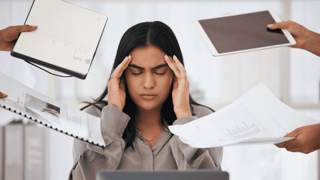 A stressed young woman sits at a desk surrounded by hands offering her various documents and electronic devices, overwhelming her.