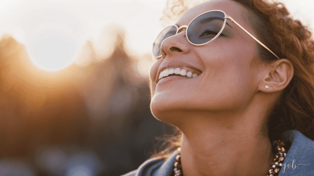 A joyful woman with curly hair, wearing sunglasses, smiles brightly in the warm sunlight.