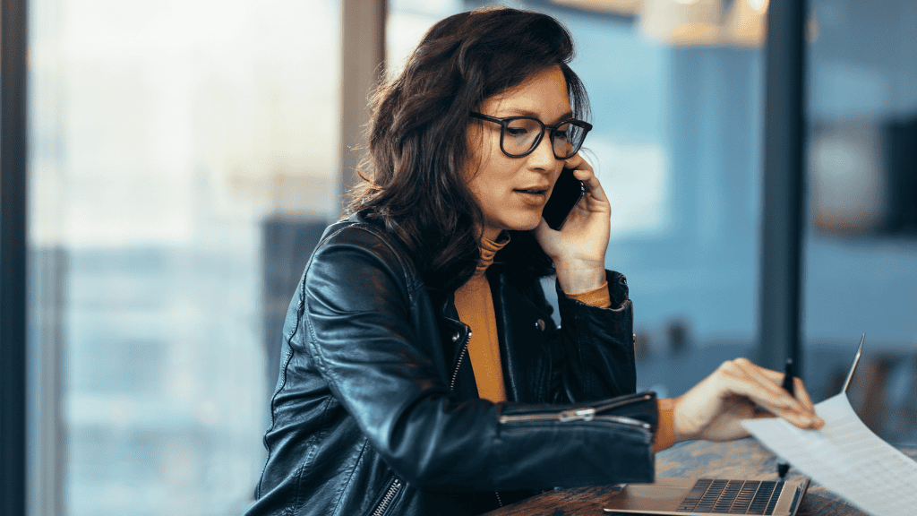 A woman in a leather jacket and glasses is on a phone call while looking at notes and sitting by a window in a modern office setting.