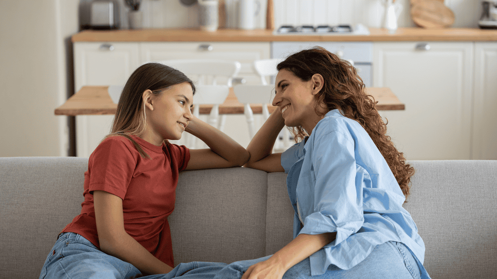 A smiling woman in a blue shirt and a teenage girl in a red t-shirt engage in a heartfelt conversation on a couch