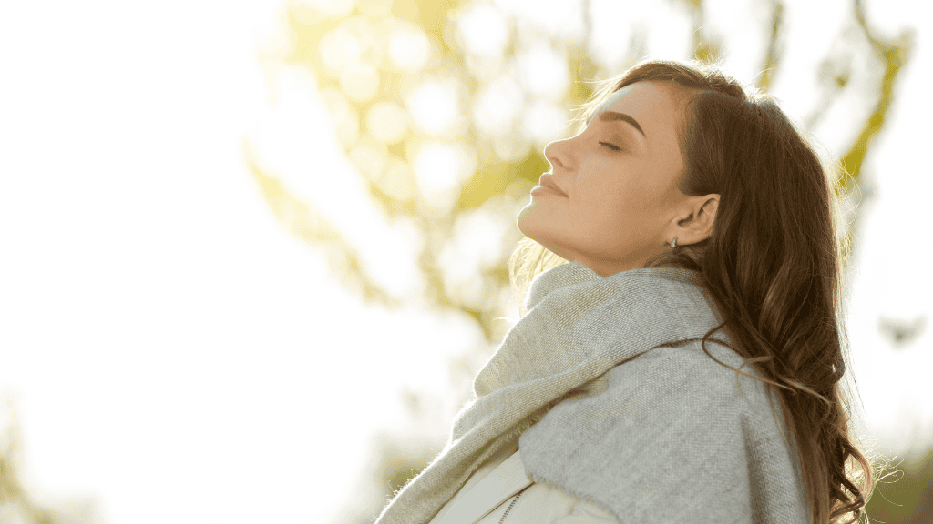 A woman with closed eyes, enveloped in sunlight, enjoys a peaceful moment outdoors, wrapped in a cozy gray scarf.