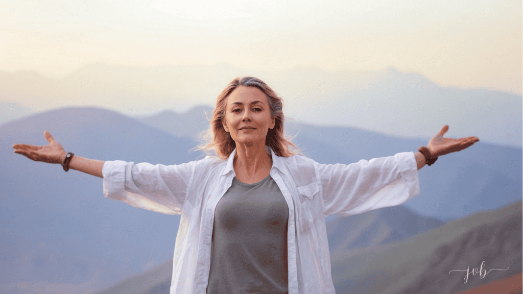 A mature woman stands with arms outstretched, embracing the vast mountain landscape at sunset.