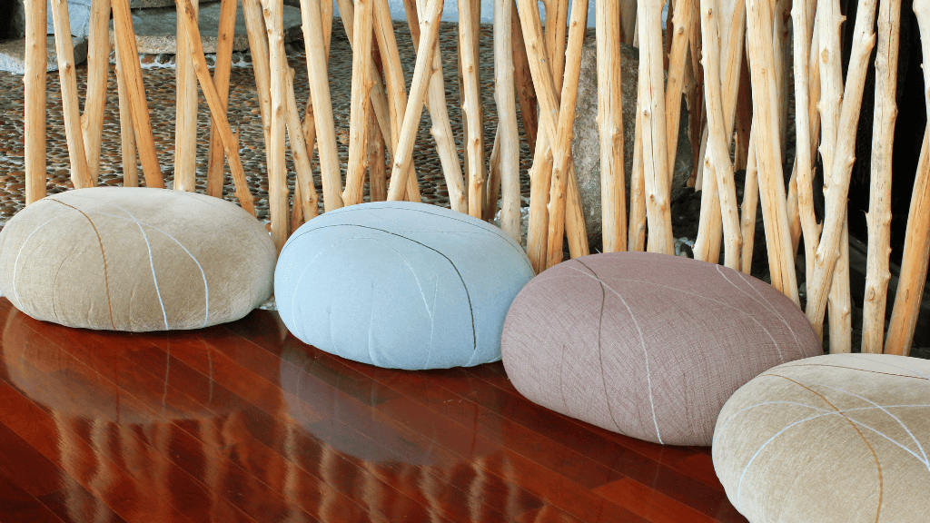 Four meditation cushions in neutral tones placed in front of a rustic wooden fence indoors, reflecting a serene and mindful environment.