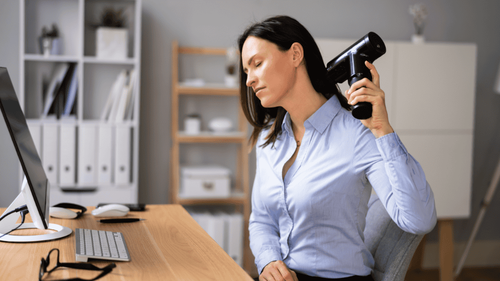 Businesswoman using a handheld deep tissue massage gun on her shoulder at her office desk