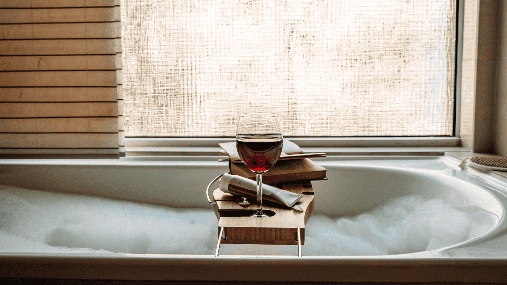 Bath caddy with a glass of red wine, a book, and bubble bath in a sunlit bathroom.