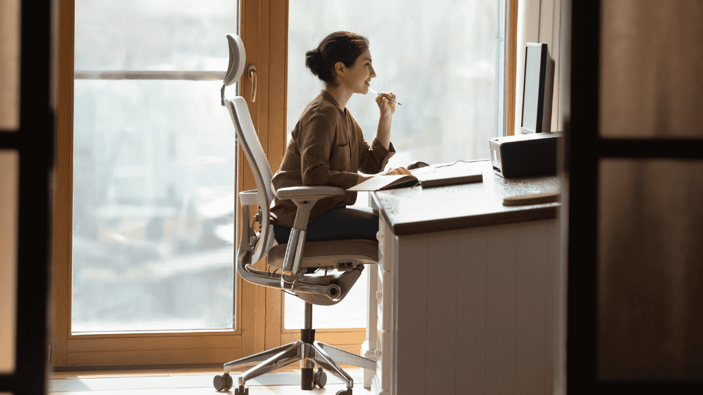 Woman thoughtfully working at a computer desk with ergonomic chair in a sunny office.