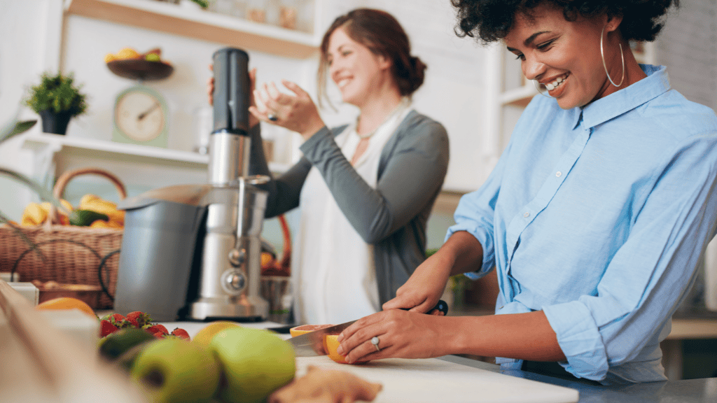 Two women preparing healthy drinks with a juicer in a sunny kitchen.