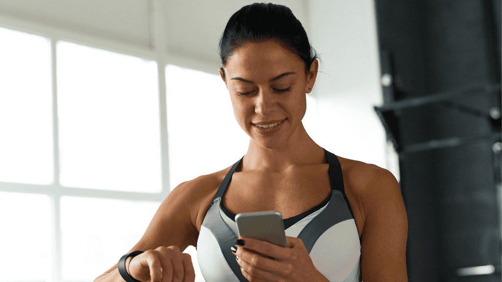 Fit woman checking fitness tracker while using smartphone in a bright gym.