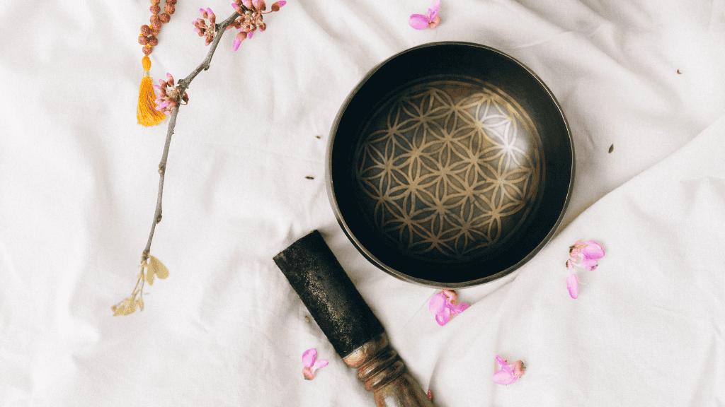 A singing bowl with a patterned interior, surrounded by cherry blossoms and a mallet, on a white fabric background.