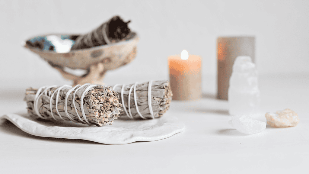 A close-up of sage bundles, a lit candle, and various crystals arranged neatly on a white surface, symbolizing elements used in spiritual practices.