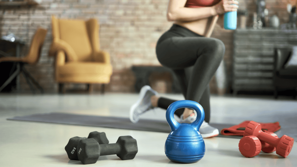 A home fitness setup with a woman squatting, holding a water bottle, and workout gear like kettlebells and dumbbells in the foreground.