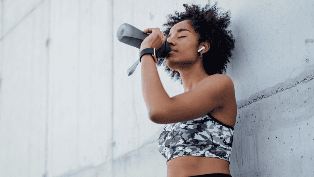 A young woman in a camo sports bra and black wrist wraps drinks from a water bottle, resting against a concrete wall during a workout break, with wireless earphones in her ears.