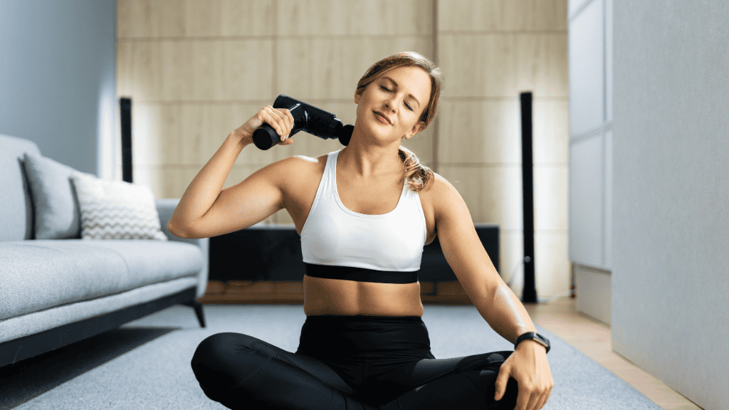 A woman in white and orange workout attire utilizes a deep tissue massage gun on her leg while sitting on a yoga mat in a serene indoor setting.