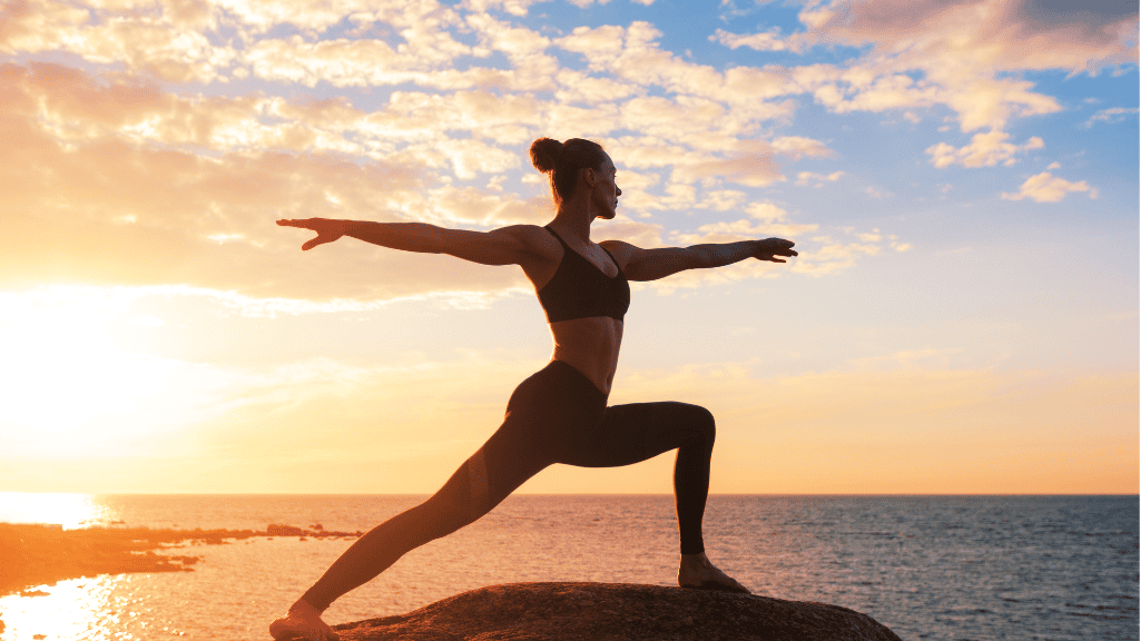 A woman practices yoga on a rocky shoreline at sunset, gracefully holding a warrior pose against a backdrop of a golden sky and calm sea.