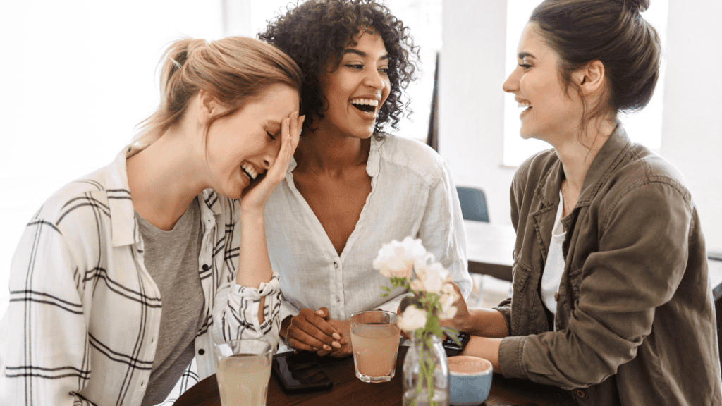 Three women laughing joyfully together at a table, sharing a light-hearted moment over drinks, embodying the strength of supportive friendships.