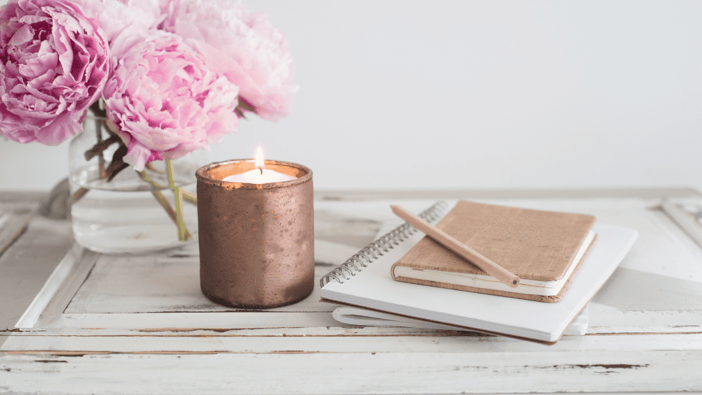 A peaceful workspace with vibrant pink peonies, a glowing candle, and journals on an antique white wooden table.