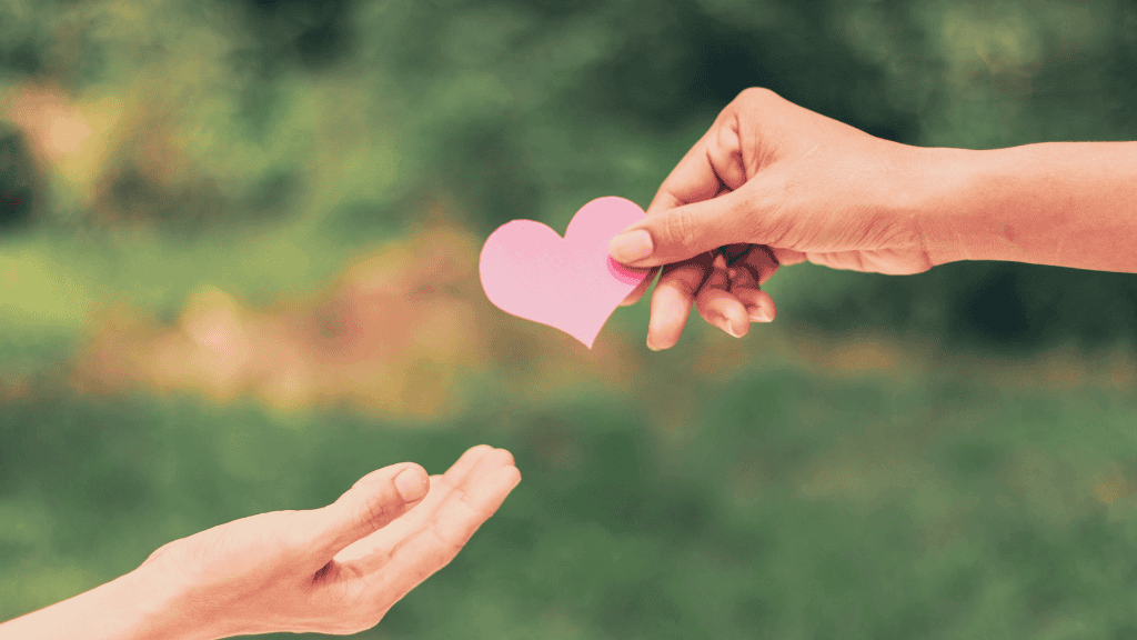 Two hands holding a pink paper heart against a blurred green background.