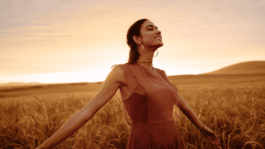 A young woman with closed eyes and a peaceful smile stands in a wheat field at sunset.