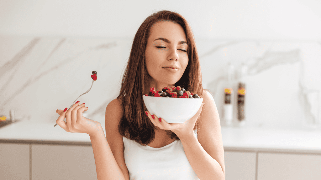 A young woman with eyes closed savoring a bowl of mixed berries, with spice jars in the background.