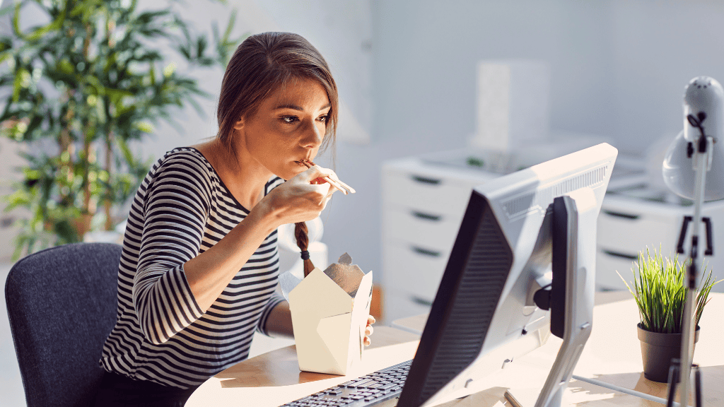 A busy woman distractedly eating lunch while working at her office desk.