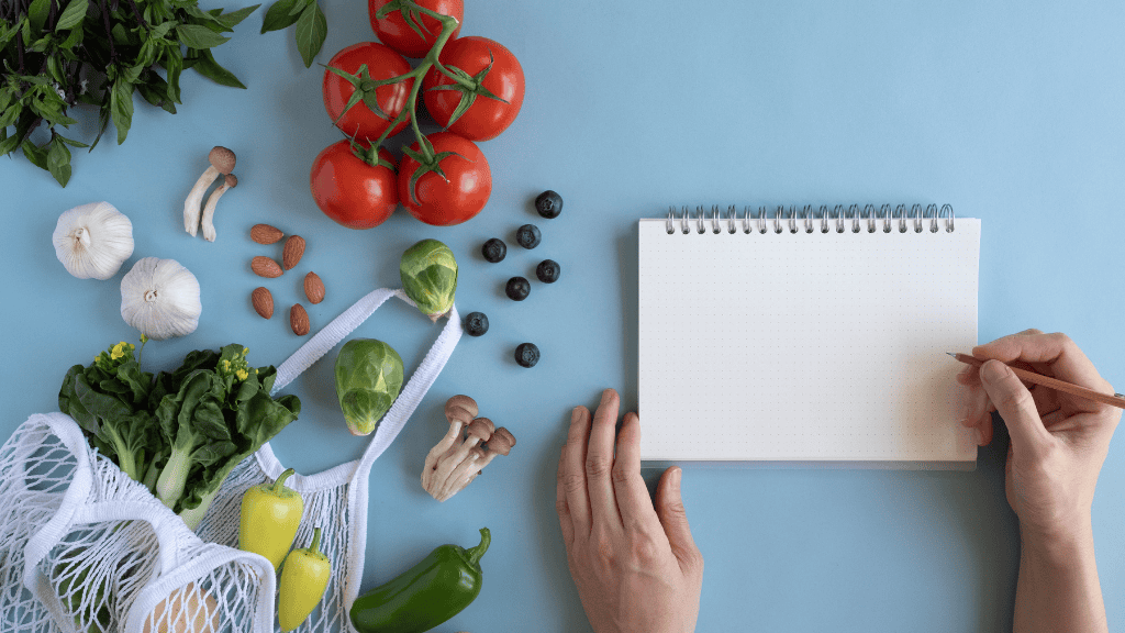 A person's hands writing in a notebook beside fresh, colorful vegetables and fruits.