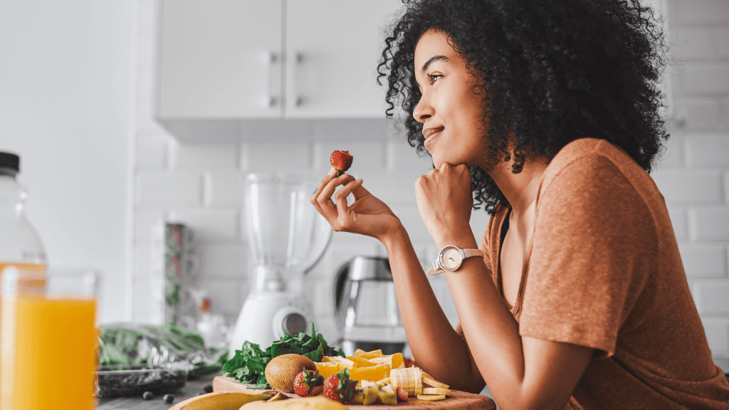 A young woman mindfully eating a strawberry while preparing a colorful meal in her kitchen.