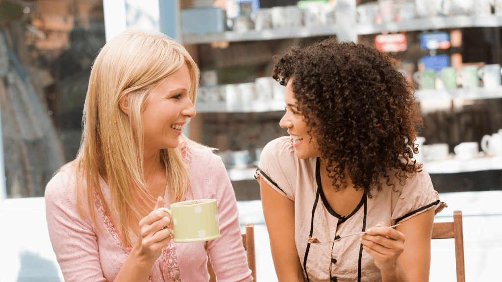 Two smiling women sitting together, one with blonde hair and one with curly brown hair, appearing to have a friendly conversation over coffee or tea.