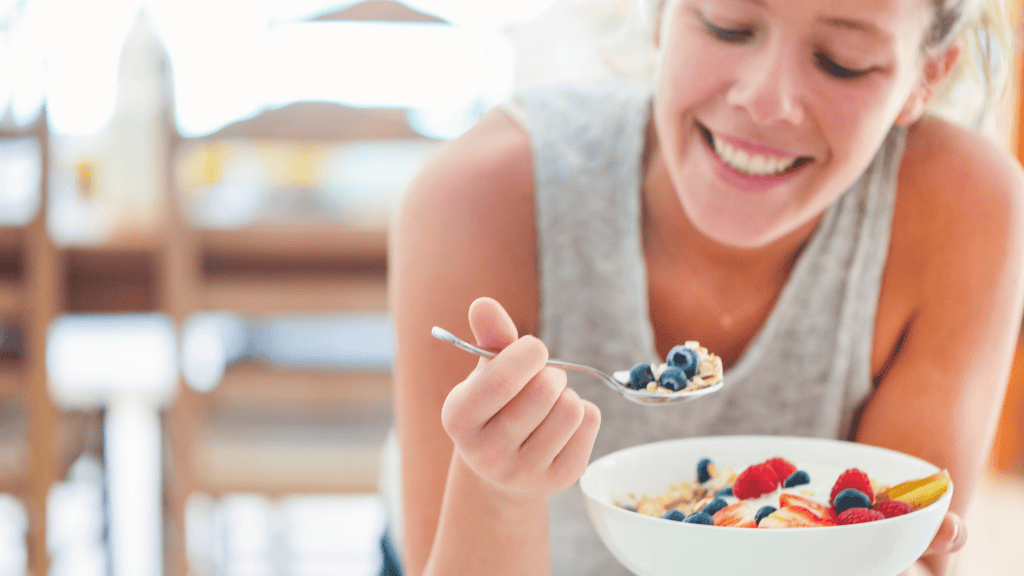 A smiling woman eating a bowl of yogurt and fresh berries, sitting at a table with sunlight coming through a window.