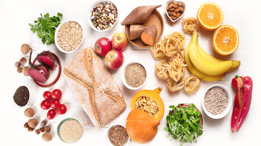 A diverse selection of carb-rich foods including whole grains, fruits, and vegetables spread out on a white surface.