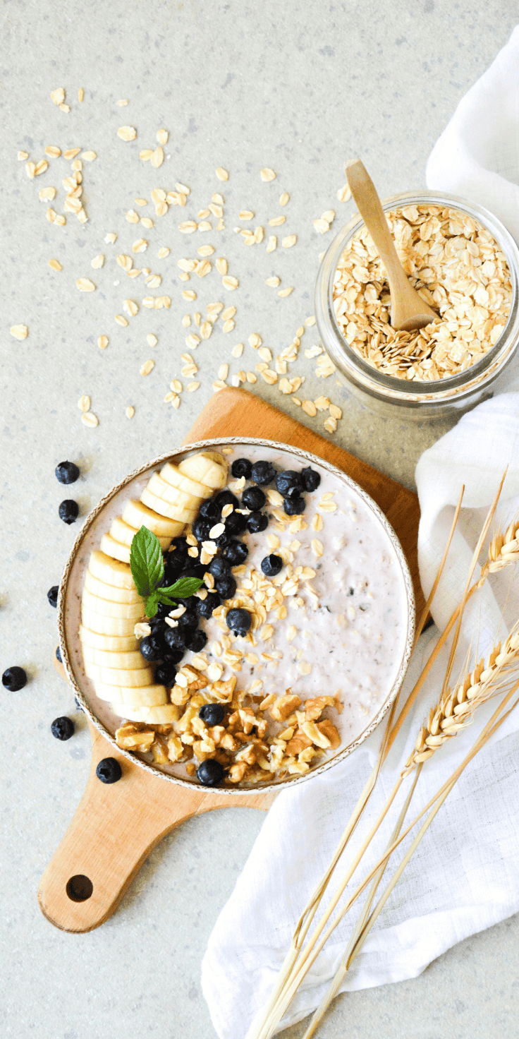 A bowl of oatmeal topped with banana slices, blueberries, walnuts, and a mint leaf sits on a wooden board next to a jar of overnight oats and scattered oats, with a white cloth and wheat stalks nearby.