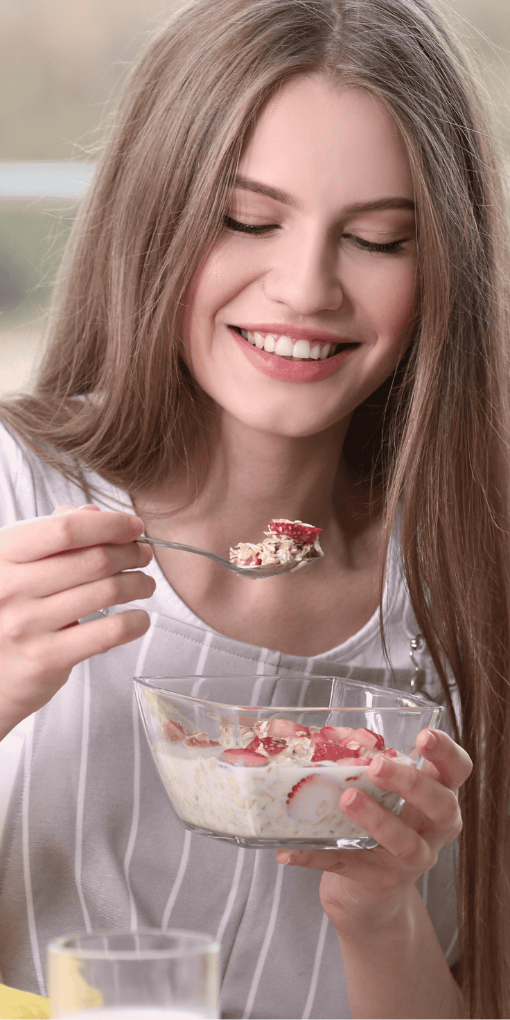 A young woman with long brown hair smiles while enjoying a bowl of overnight oats with strawberries, eating it with a spoon.