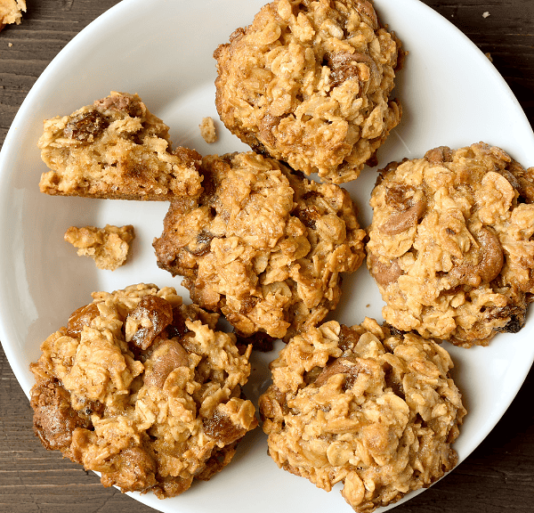 A plate of breakfast cookies sits on a wooden table, perfect for on-the-go mornings. Some oatmeal raisin cookies are crumbled, revealing their hearty texture. A few raisins are scattered around the plate.