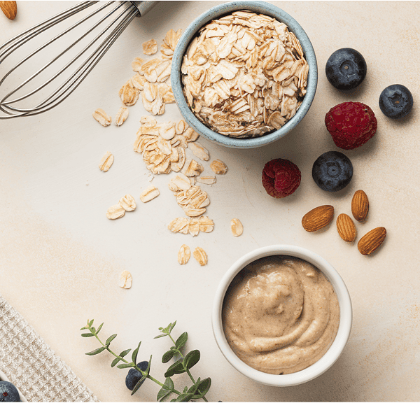 A flat lay of protein-rich ingredients on a light surface showcases oats, almonds, and berries alongside a bowl of creamy nut butter, a whisk, and a small plant. This vibrant arrangement suggests preparation for a healthy breakfast to energize your mornings.