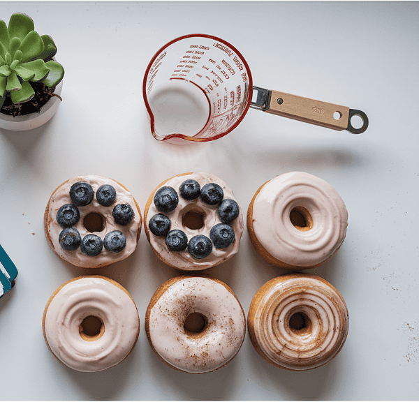 A floral notebook and a small potted succulent sit on a table beside six protein donuts with icing, two topped with blueberries. Nearby, a measuring cup with a wooden handle holds white liquid. The table embodies a clean, minimalist design perfect for jotting down new protein donut recipes.