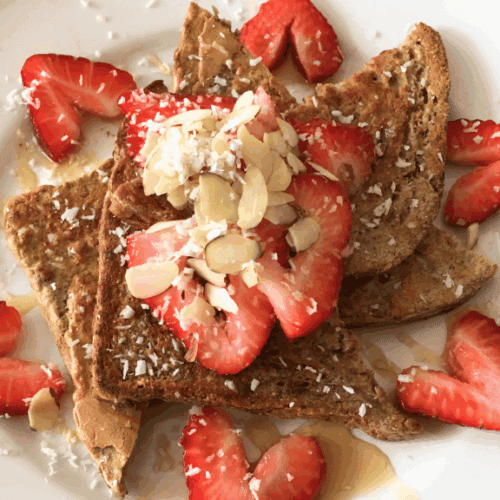 Three slices of French toast topped with sliced strawberries, almond flakes, shredded coconut, and a drizzle of syrup, served on a white plate with extra strawberry slices—a delicious and healthy meal for any time of day.