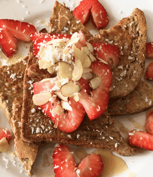 Three slices of French toast topped with sliced strawberries, almond flakes, shredded coconut, and a drizzle of syrup, served on a white plate with extra strawberry slices—a delicious and healthy meal for any time of day.