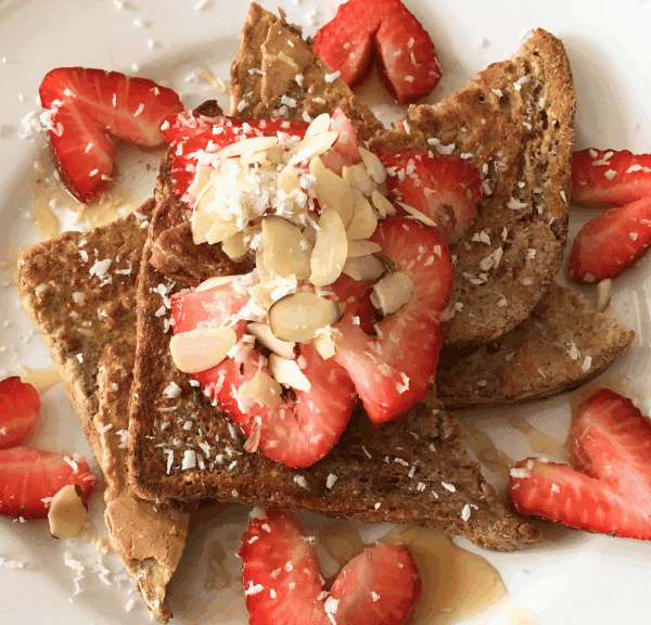 Three slices of French toast topped with sliced strawberries, almond flakes, shredded coconut, and a drizzle of syrup, served on a white plate with extra strawberry slices—a delicious and healthy meal for any time of day.