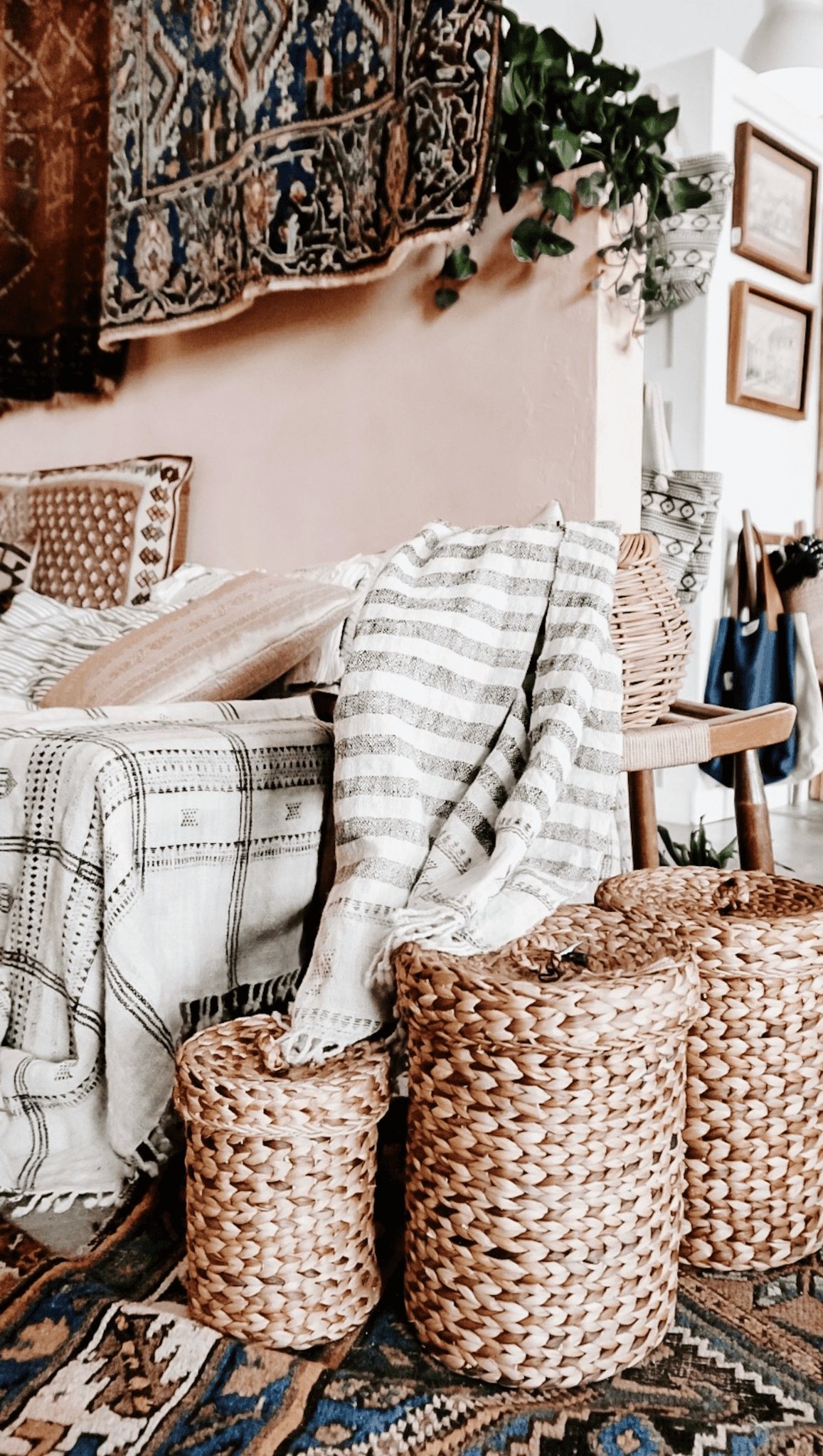 A cozy bohemian living space doubles as a meditation room, with a patterned rug, woven baskets, a striped throw blanket on the sofa, and textured pillows. Decorative wall hangings and green plants create a warm, inviting atmosphere.