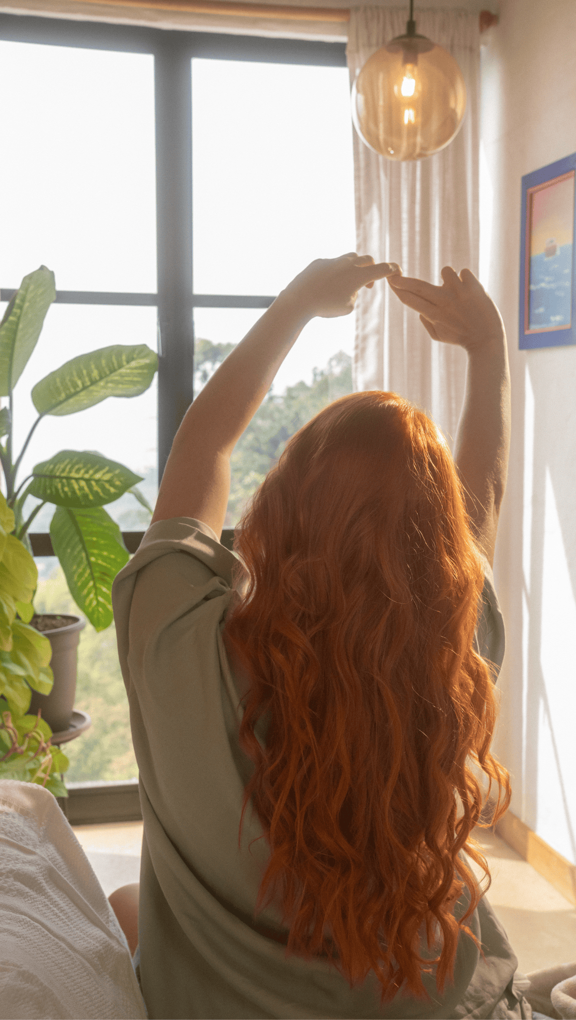 A person with long, wavy red hair stretches with arms raised while sitting on a bed in a sunlit room, enjoying their morning rituals near a window with plants and framed art on the wall.