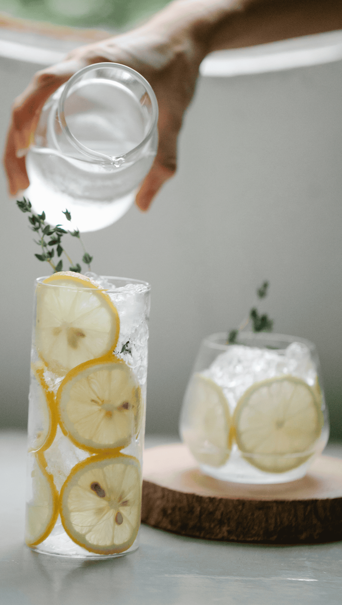 A hand pours water from a small pitcher into a tall glass filled with ice, lemon slices, and thyme—a refreshing boost metabolism drink for your morning rituals. Another glass rests on a wooden coaster in the background.