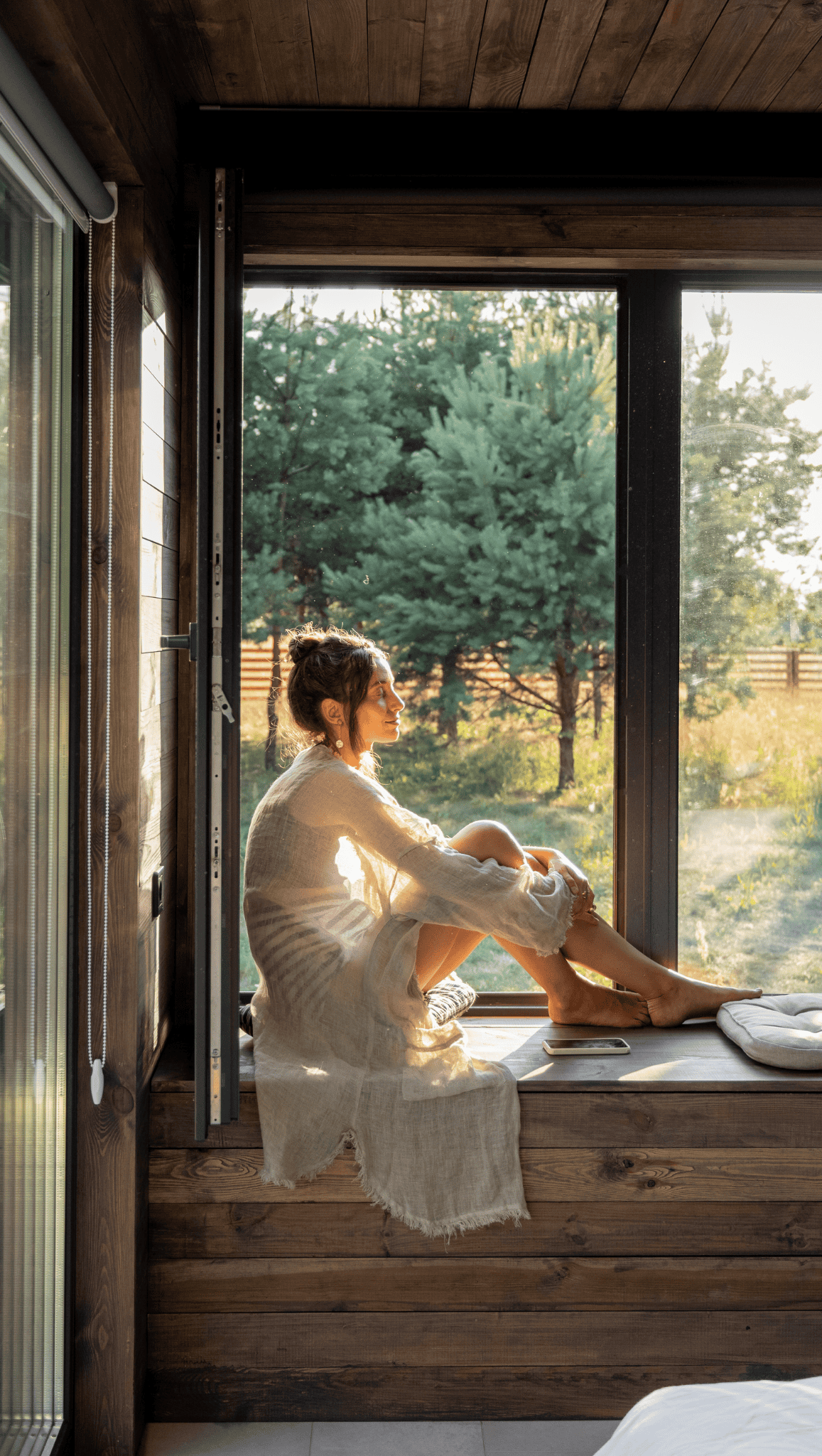 A woman in a light, loose dress sits on a windowsill in a wooden cabin, bathed in warm sunlight. As part of her morning rituals, she gazes thoughtfully outside at green trees and a sunlit garden to boost metabolism and start her day.
