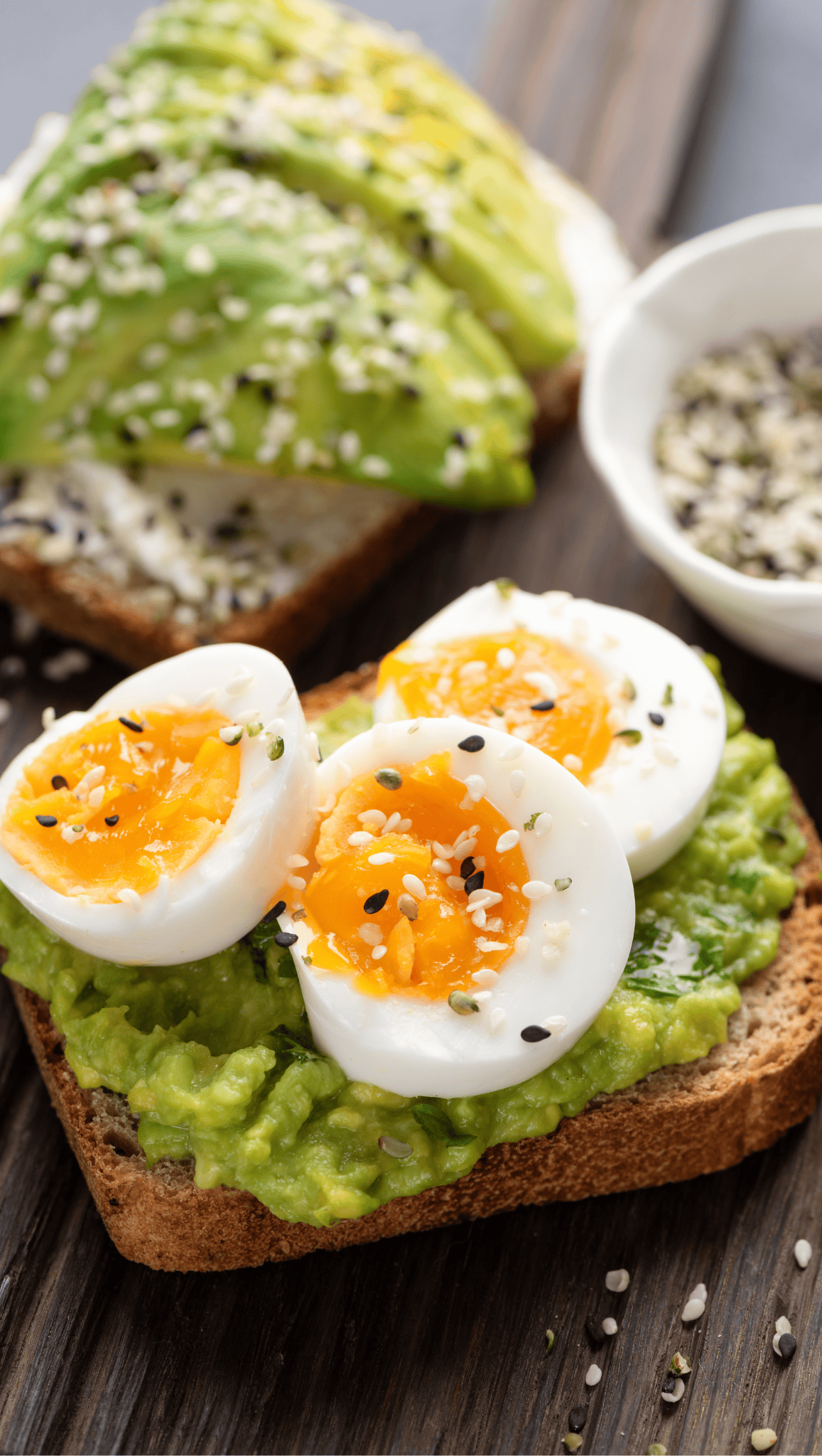 Elevate your morning rituals with two slices of whole grain toast topped with creamy mashed avocado; one slice features soft-boiled eggs, both sprinkled with sesame seeds to help boost metabolism. A small bowl of seeds rests nearby on a wooden surface.