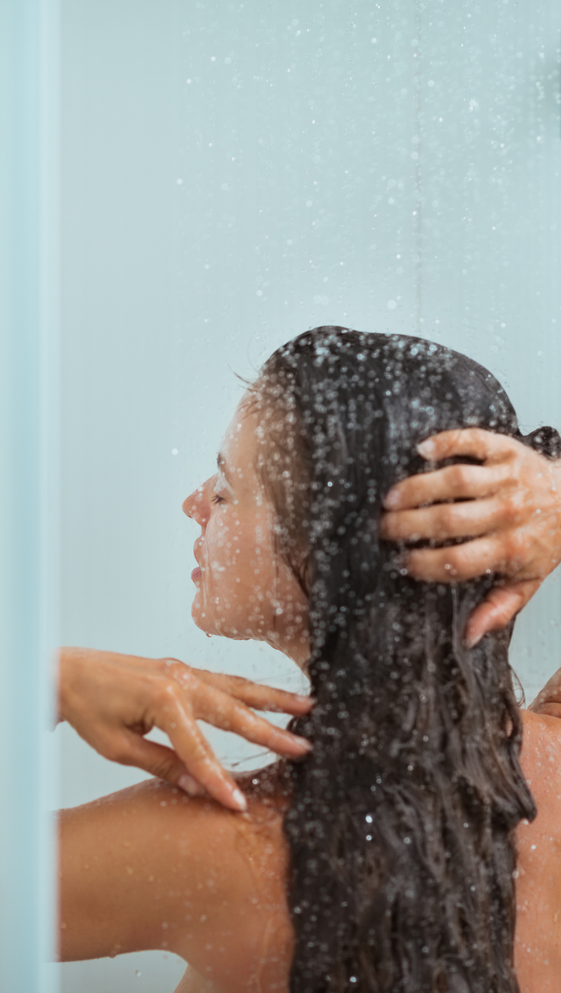 As part of her morning rituals, a woman with long dark hair takes a shower, running her hands through her hair under falling water, with droplets visible on the glass.