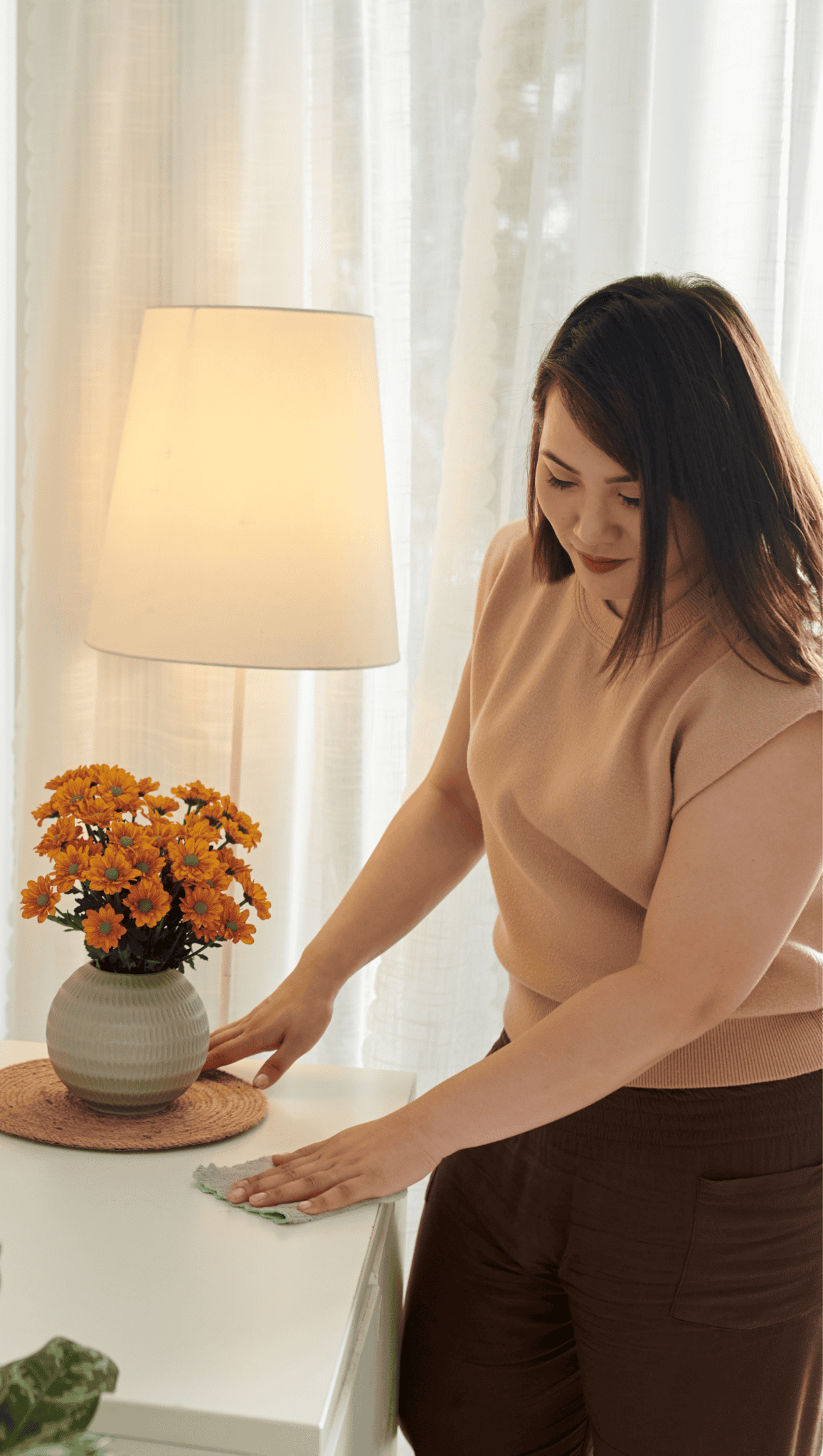 A woman wipes a white table near a lamp and a vase of orange flowers, creating a calming atmosphere perfect for meditation spaces, with sheer curtains in the background.