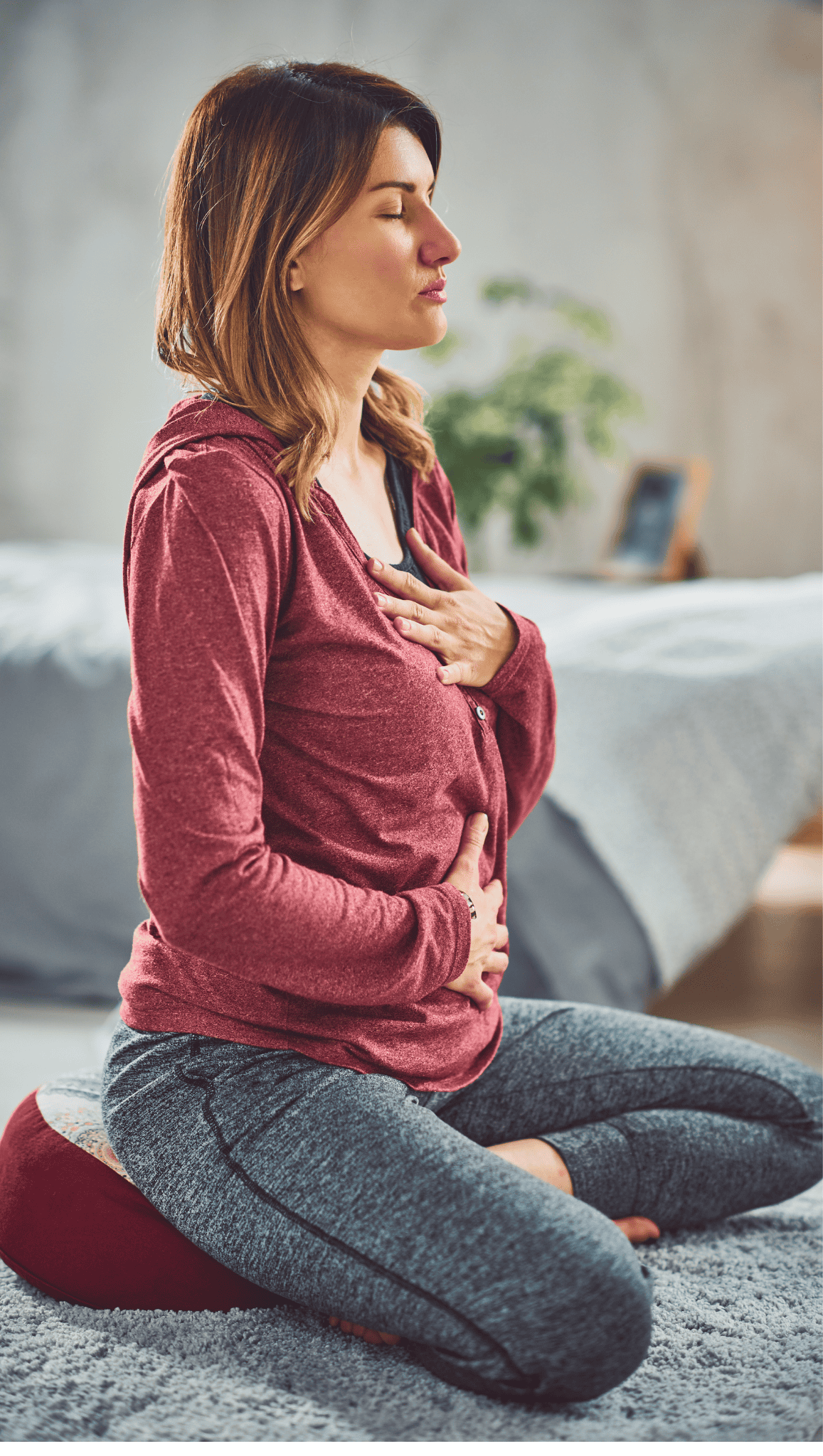 A woman in a red hoodie and gray sweatpants sits cross-legged on a cushion indoors, eyes closed, with one hand on her chest and one on her abdomen, practicing deep breathing as part of her morning rituals to boost metabolism.