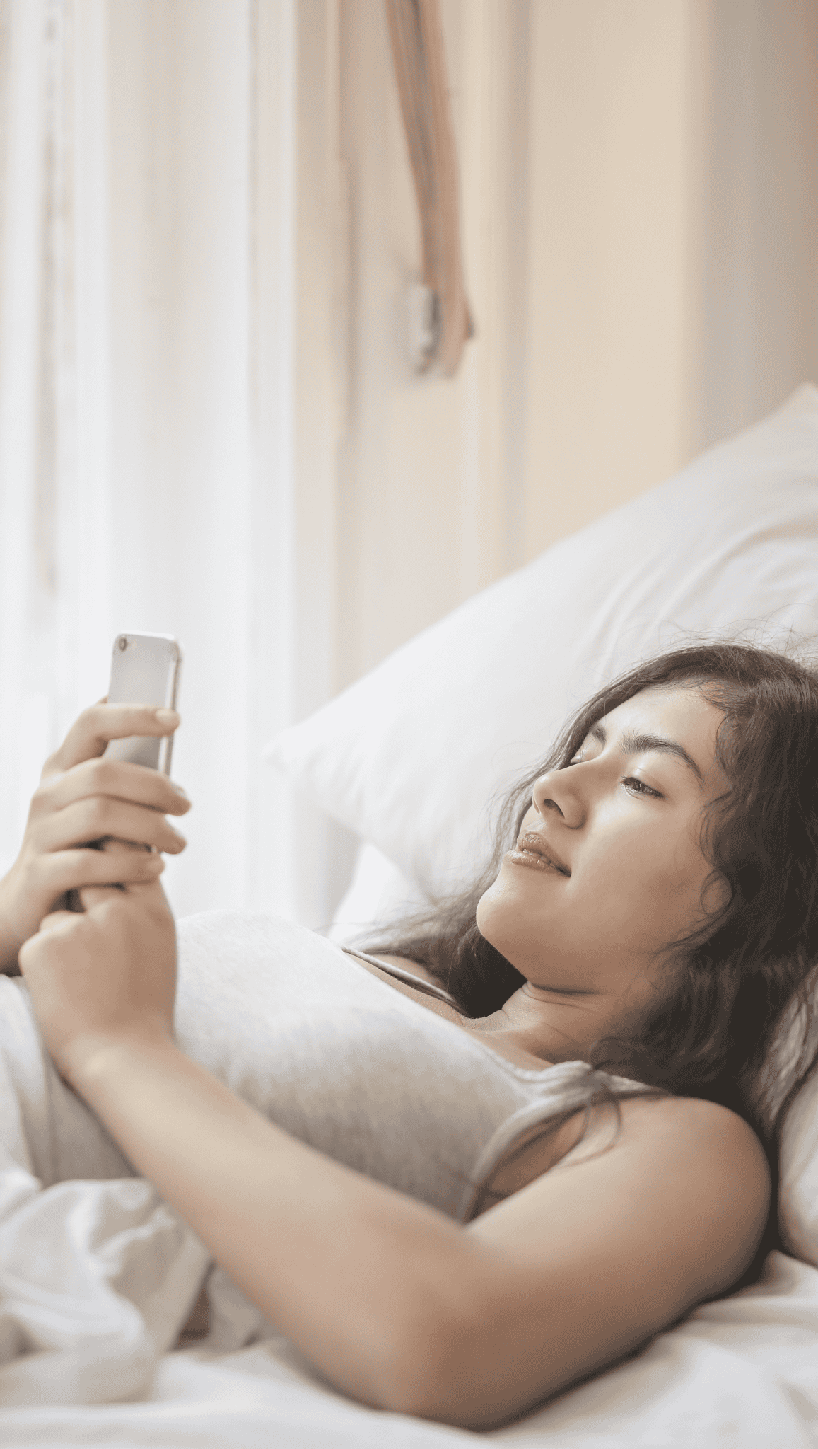 A young woman with long dark hair is lying in bed, propped up on pillows, holding and looking at her smartphone with a relaxed expression—enjoying her morning rituals to boost metabolism as soft natural light comes through a nearby window.