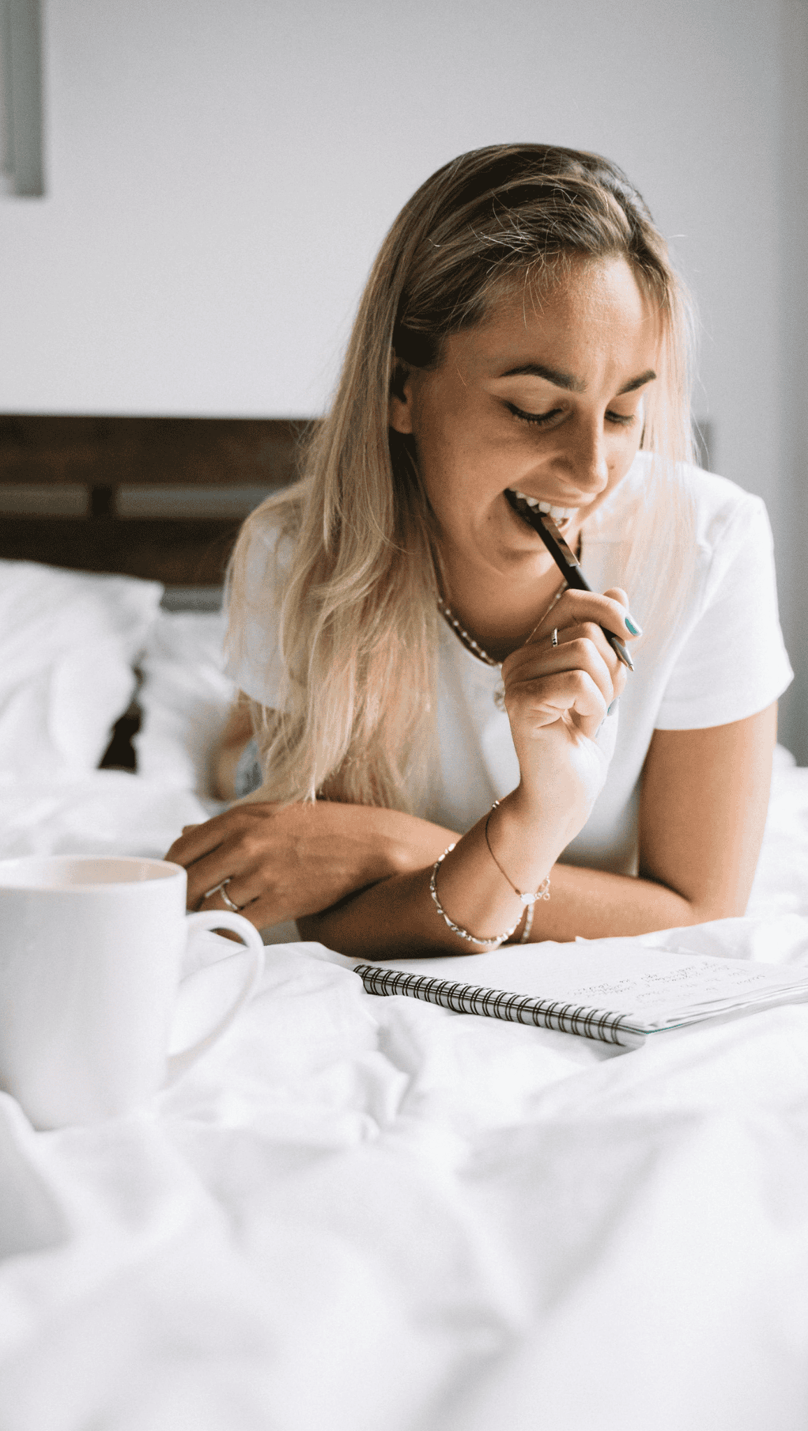 A woman with long blonde hair lies on a bed, smiling while writing in a spiral notebook as part of her morning rituals. A white mug rests nearby, adding to the relaxed and bright atmosphere.