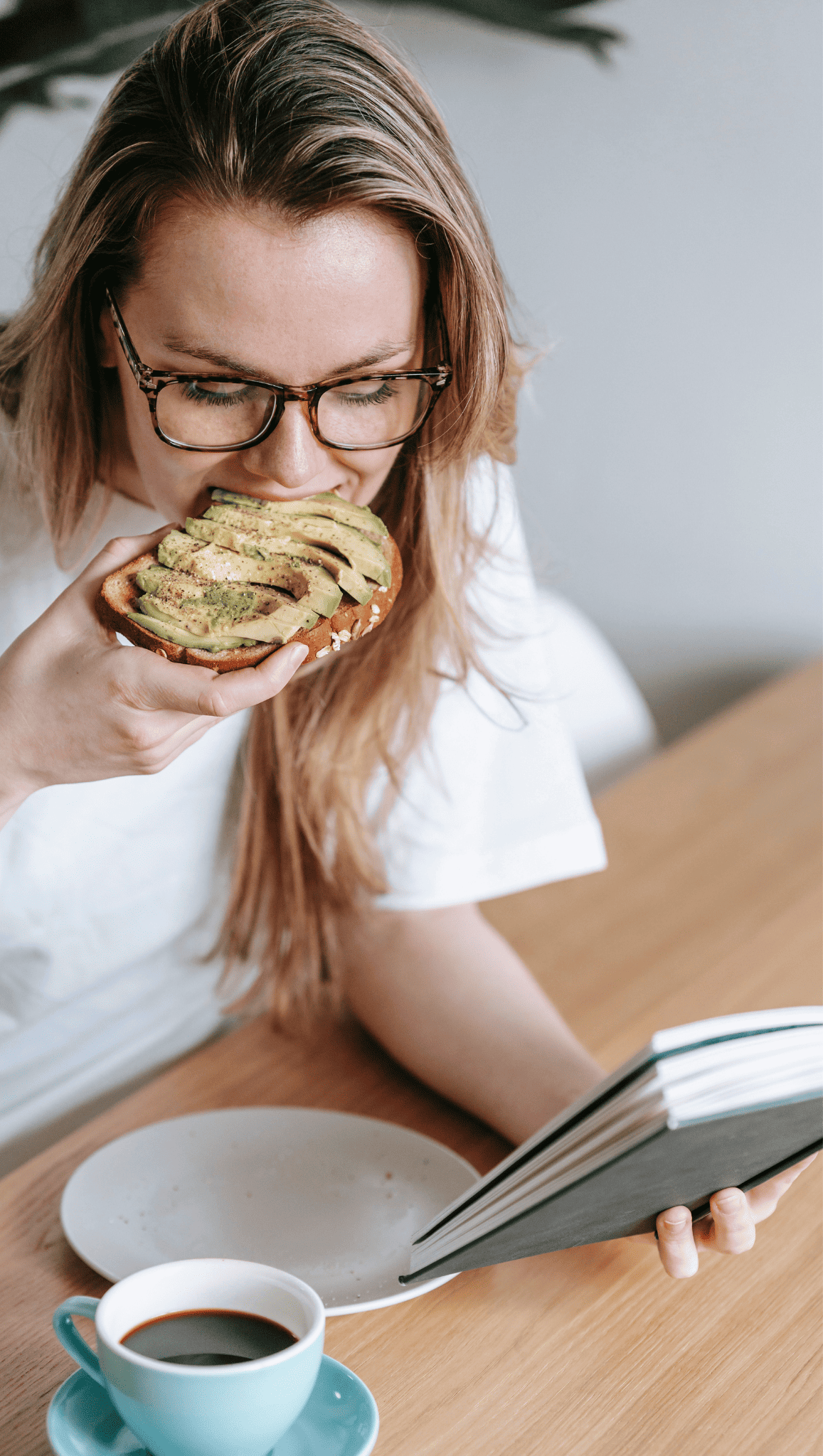 Engaged in her morning ritual, a woman with long blonde hair and glasses eats avocado toast while reading a book at a wooden table. A cup of coffee and an empty plate sit in front of her, helping to boost metabolism for the day ahead.