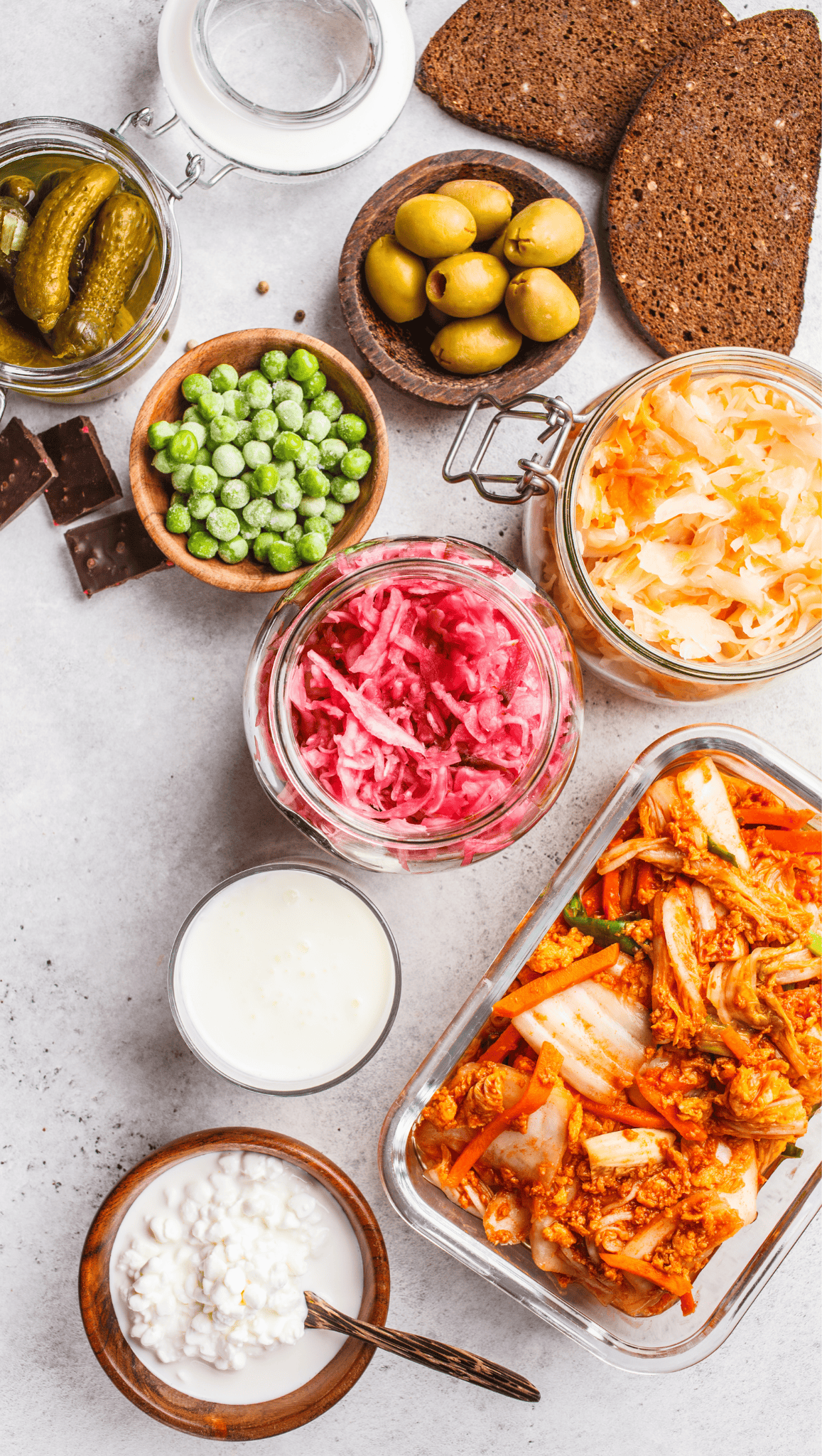 A variety of fermented and probiotic foods are displayed on a light surface, perfect for morning rituals to boost metabolism. Included are kimchi, sauerkraut, pickles, olives, cottage cheese, yogurt, rye bread, green peas, and dark chocolate.