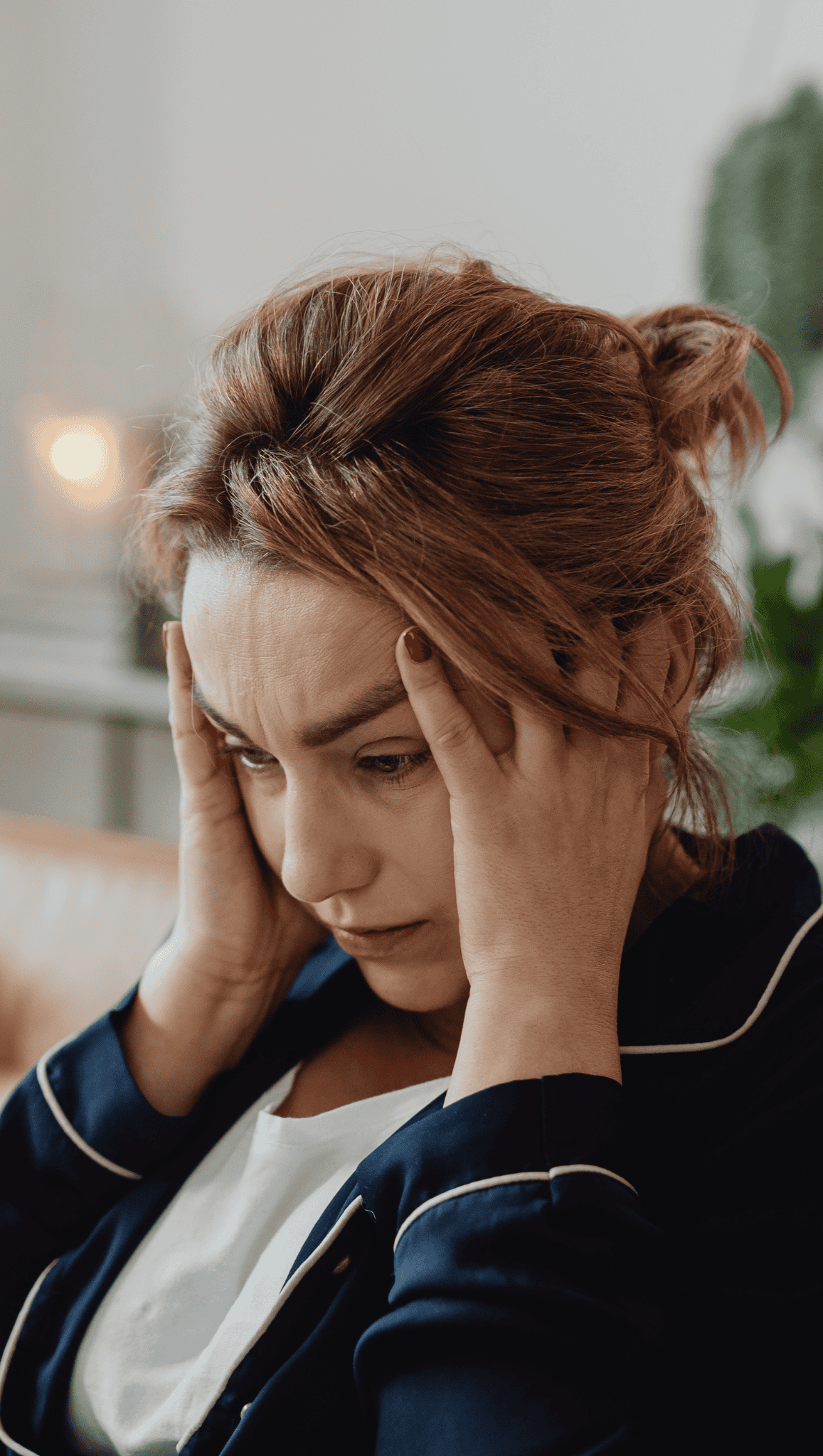 A woman with light brown hair, wearing a dark top, sits indoors holding her head with both hands and appears stressed or worried—perhaps overwhelmed by trying new morning rituals to boost metabolism.